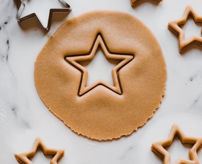 A rolled-out sheet of cookie dough with star shapes cut out using cookie cutters. The smooth, golden-brown dough has a large central star and smaller stars around it, ready for the stained glass cookie effect. This classic holiday cookie preparation hints at a festive treat with a beautiful translucent design.