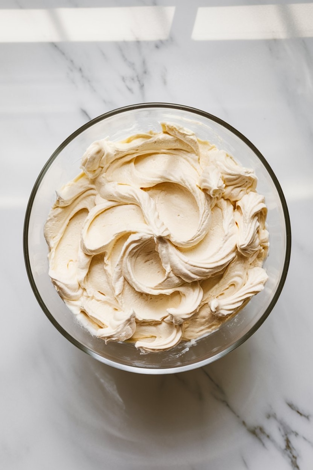 A bowl of creamy vanilla frosting in a clear glass bowl on a marble countertop, under natural lighting.