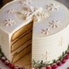 Festive winter-themed layer cake with smooth white frosting, decorated with intricate snowflake designs and a dusting of powdered sugar. A slice is cut to reveal the even layers of sponge cake and creamy filling inside. The cake is garnished at the base with fresh cranberries and rosemary sprigs, adding a pop of color and holiday charm to this elegant Christmas dessert.