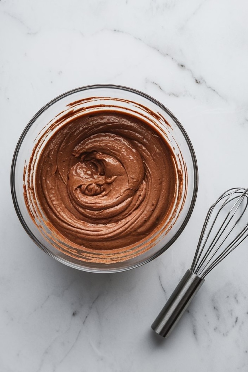 A bowl of silky, chocolate cupcake batter, freshly mixed and swirled to a smooth consistency, ready for baking. The rich, creamy texture of the batter, with its deep brown color, highlights the decadence of the chocolate. A stainless steel whisk sits beside the bowl on the marble countertop, adding to the scene of a cozy baking session perfect for creating winter treats.