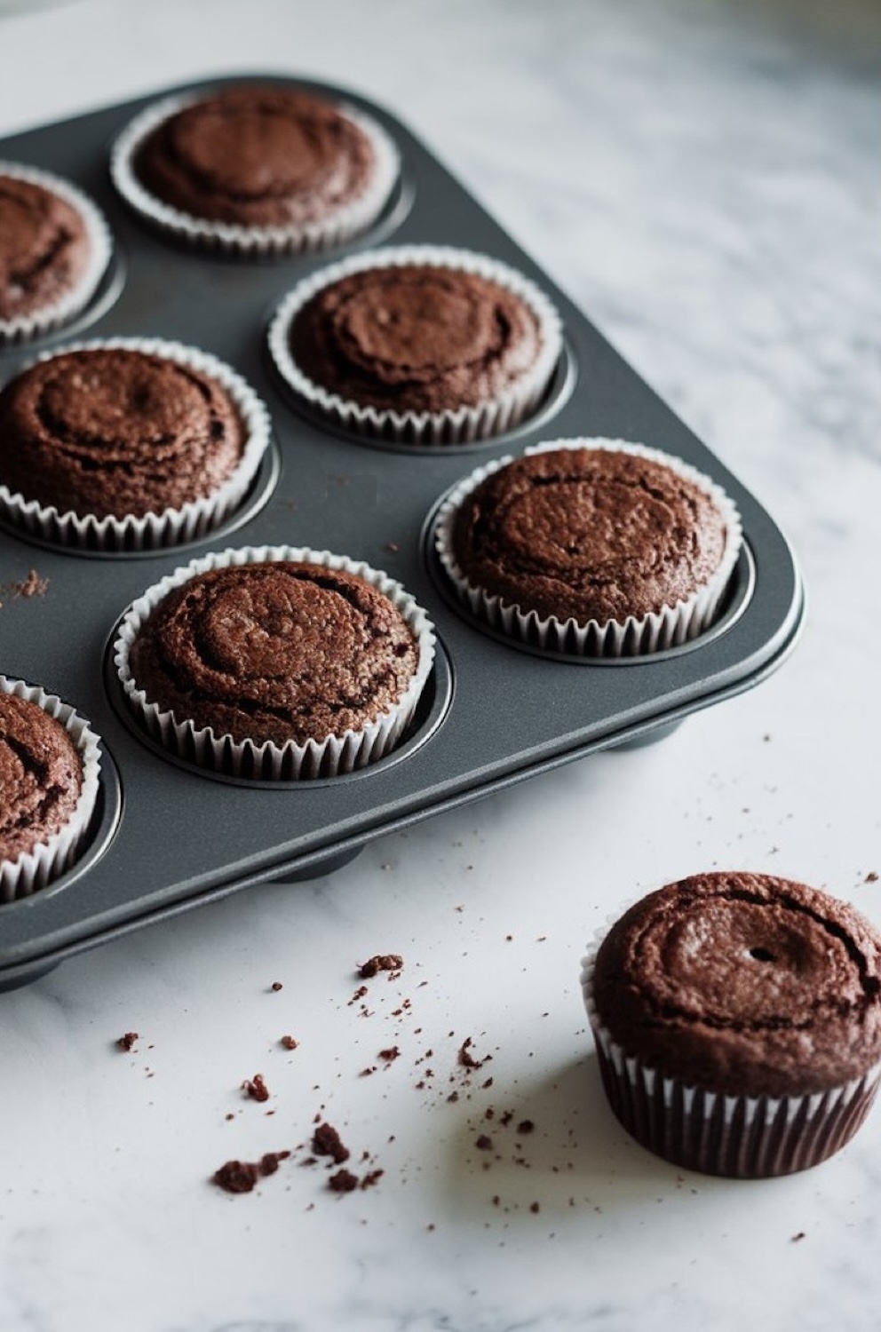 Freshly baked chocolate cupcakes in a dark muffin tin, with one cupcake set beside it on a marble countertop. The cupcakes have a rich, fluffy texture, with a slightly cracked top that reveals their moist interior. Loose crumbs scattered on the surface add a homemade, rustic touch, making them look irresistibly delicious and ready for frosting.