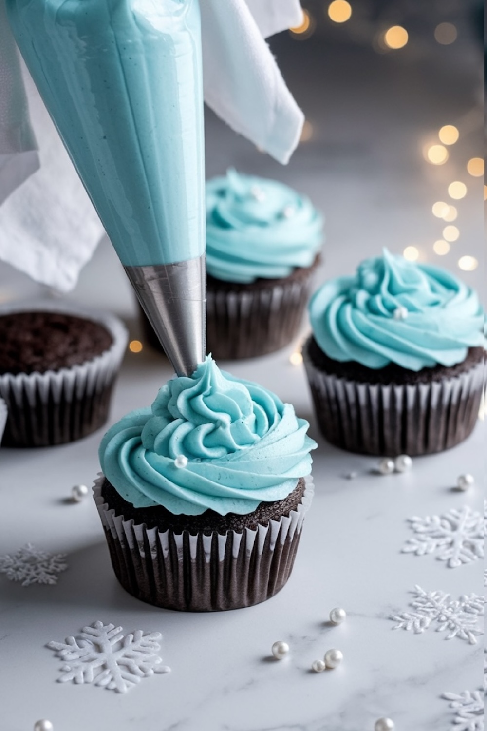 A close-up of chocolate cupcakes being decorated with swirls of pastel blue frosting, creating a wintery, frozen-inspired look. The smooth, creamy frosting is applied with a piping bag, adding texture and an elegant touch. Small white pearl sprinkles and delicate snowflake decorations on the marble surface enhance the winter wonderland theme, while soft fairy lights in the background create a cozy, festive atmosphere.