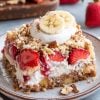 A full plate of banana split dump cake, topped with fresh banana slices, whipped cream, strawberries, and crushed pecans. The background includes a rustic cutting board with whole strawberries and bananas.
