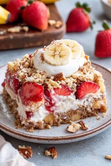 A full plate of banana split dump cake, topped with fresh banana slices, whipped cream, strawberries, and crushed pecans. The background includes a rustic cutting board with whole strawberries and bananas.