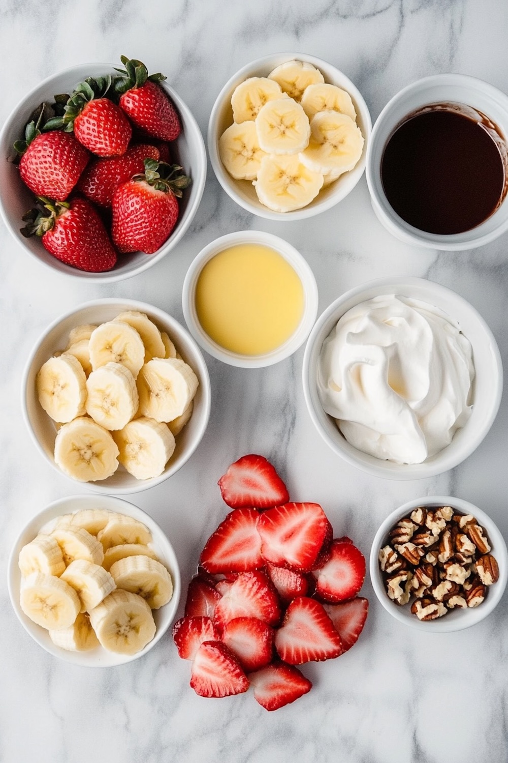 Flat-lay of dessert ingredients, including fresh strawberries, sliced bananas, whipped cream, chocolate sauce, chopped pecans, and a yellow custard-like sauce, all arranged in white bowls on a marble countertop.
