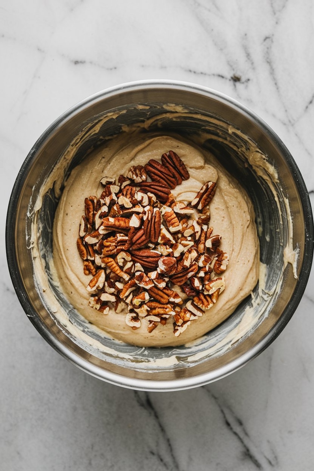 A steel mixing bowl containing smooth cake batter with chopped pecans generously sprinkled on top, ready to be folded in. The marbled countertop provides a clean, aesthetic background for the baking process.