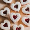 Heart-shaped Linzer cookies filled with cherry jam, dusted with powdered sugar, arranged on a wooden board for presentation.