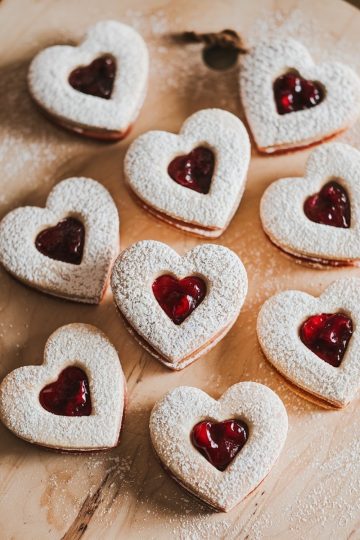 Heart-shaped Linzer cookies filled with cherry jam, dusted with powdered sugar, arranged on a wooden board for presentation.