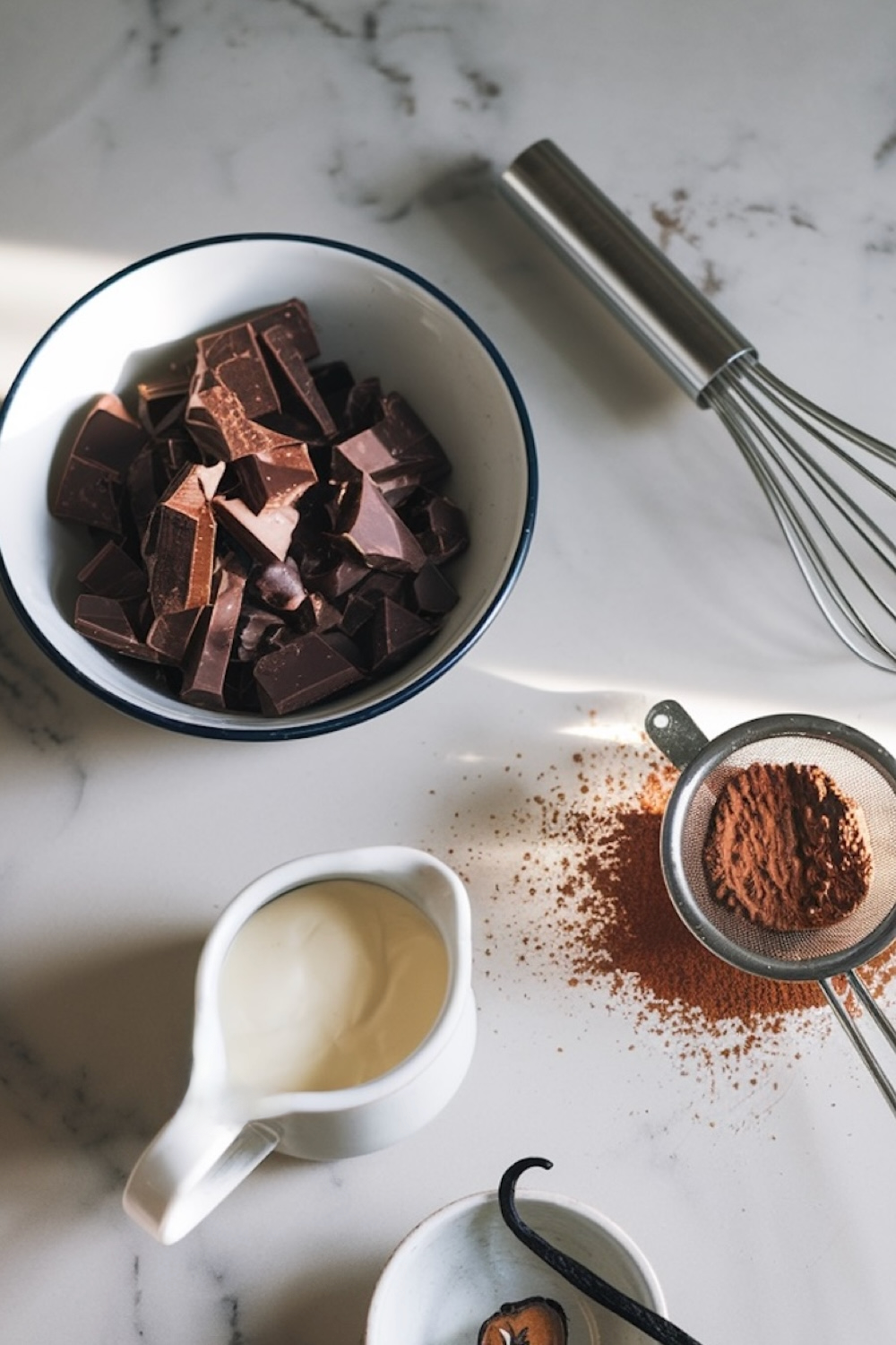 Ingredients for chocolate mousse on a marble surface, including chopped dark chocolate in a bowl, cocoa powder, heavy cream in a pitcher, and a whisk.
