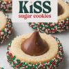 Close-up of a Christmas kiss sugar cookie with festive holiday sprinkles. The sugar cookie is topped with a classic chocolate Hershey’s kiss and surrounded by red, green, and white sprinkles for a festive touch. Above the cookie, bold text reads “Christmas Kiss Sugar Cookies” in holiday-inspired colors, perfect for a holiday baking or Christmas dessert idea.