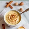 Bowl of thick homemade eggnog with cinnamon dusting and a swirl pattern, surrounded by whole nutmeg and cinnamon sticks, with a slice of bread spread with eggnog cream on the side.
