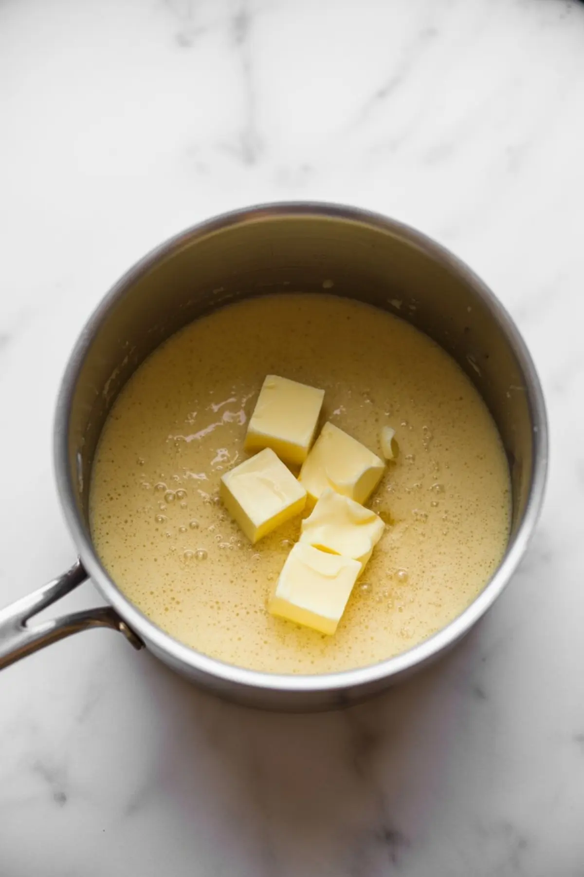 Chunks of butter melting into a warm pot of thick eggnog mixture, creating a rich, golden-yellow base in a stainless steel saucepan.