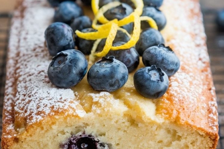 A beautifully baked lemon blueberry pound cake topped with fresh blueberries, lemon zest curls, and a dusting of powdered sugar. A text overlay reads "Lemon Blueberry Pound Cake," capturing attention with a bold, inviting font and perfect for sharing this delicious dessert recipe online.