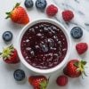A close-up of a bowl filled with thick, vibrant mixed berry jam, featuring a blend of strawberries, blueberries, and raspberries. Fresh berries are scattered around the bowl, highlighting the rich berry flavors in this homemade jam.