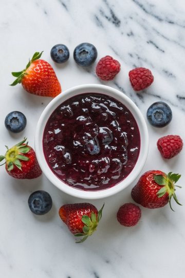 A close-up of a bowl filled with thick, vibrant mixed berry jam, featuring a blend of strawberries, blueberries, and raspberries. Fresh berries are scattered around the bowl, highlighting the rich berry flavors in this homemade jam.