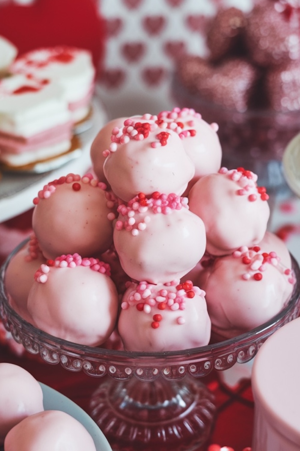 Elegant display of pink Oreo truffle balls stacked on a vintage glass dessert stand, perfect for Valentine's Day or a festive celebration. Each truffle is coated in smooth, pastel pink candy melts and topped with a sprinkle of red, pink, and white nonpareils, creating a whimsical and romantic dessert presentation. The blurred background shows heart-themed decor, enhancing the sweet and celebratory ambiance. Keywords: Valentine’s Day dessert, pink Oreo truffles, romantic party treats, festive dessert table, candy-coated Oreo balls, sprinkle-topped truffles, holiday dessert inspiration.
