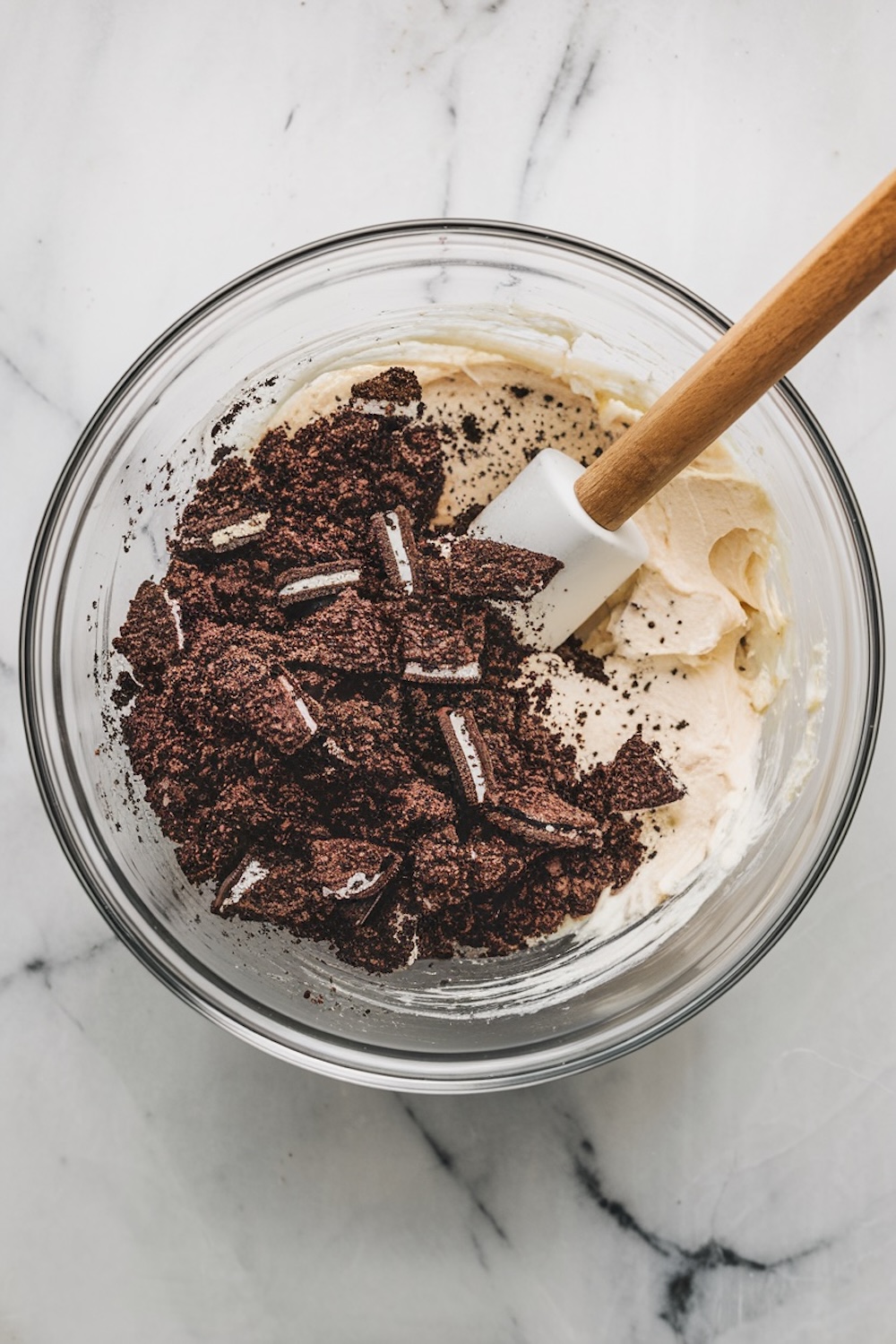  Mixing bowl with crushed Oreo cookies and cream cheese filling on a white marble countertop. The crumbled chocolate cookies are mixed into a creamy base with a silicone spatula, preparing ingredients for Oreo truffles. Keywords: Oreo truffle recipe, dessert preparation, no-bake Oreo balls, cookie and cream filling, easy dessert ideas.

