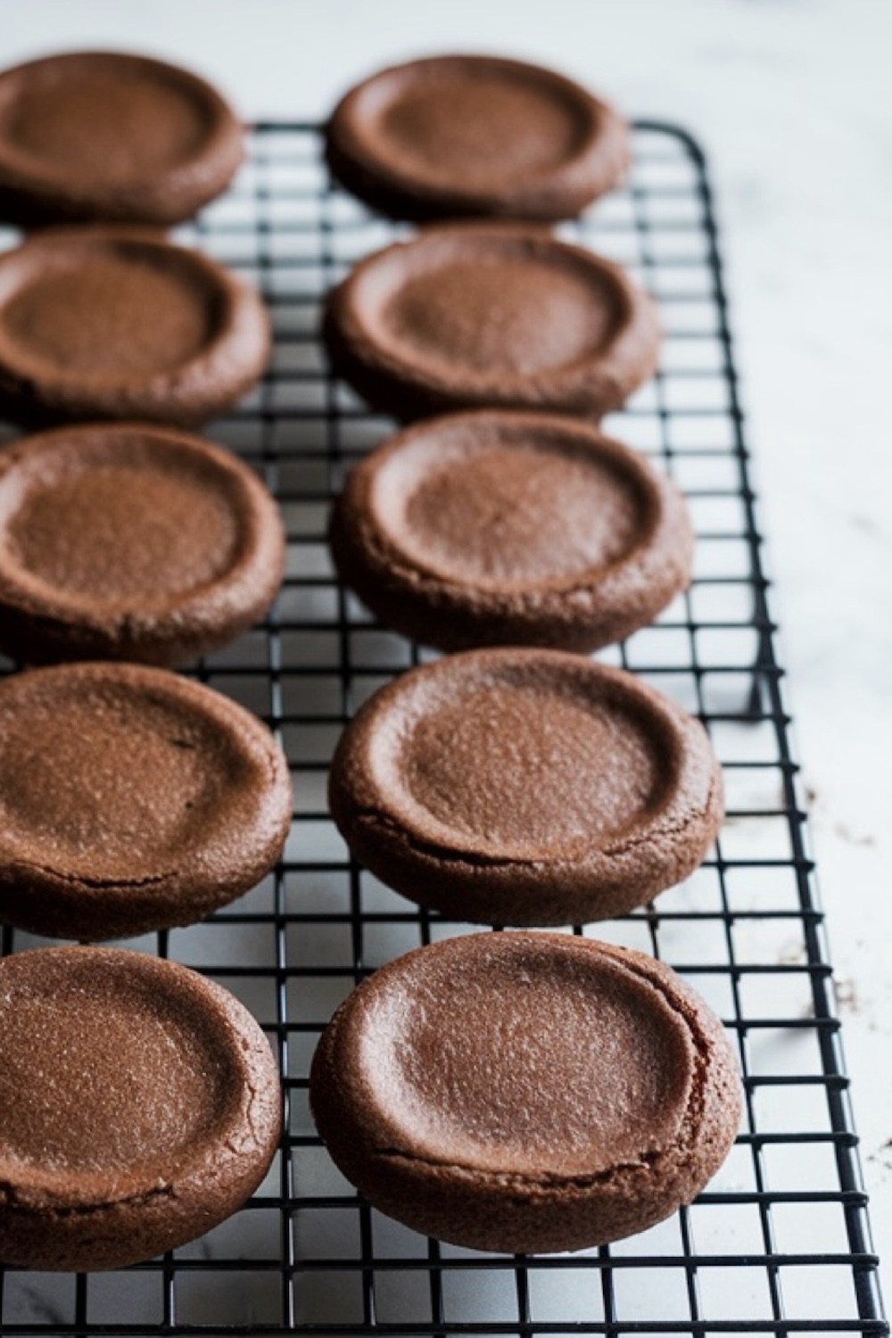 Rows of plain, freshly baked chocolate cookie bases cooling on a wire rack. These simple, rounded cookie shapes are smooth with slight indentations, ready to be coated with chocolate and transformed into peppermint bark cookies.