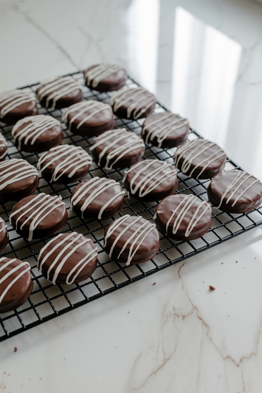 Chocolate-dipped cookies drying on a wire rack, now topped with delicate white chocolate drizzle, adding a touch of elegance and contrasting color to the rich chocolate coating.