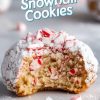 A close-up of a peppermint snowball cookie with a bite taken out, showcasing a soft, chewy interior filled with bits of crushed peppermint candy. The cookie is coated in powdered sugar, giving it a frosty, snowball-like appearance. Above, the festive text "Peppermint Snowball Cookies" is displayed in playful, holiday-themed typography, making this image perfect for capturing the cozy, seasonal feel of holiday baking.
