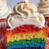 A detailed shot of a rainbow cupcake cut open, highlighting its vividly colored layers and topped with a swirl of vanilla frosting sprinkled with multi-colored confetti. The rainbow layers and smooth frosting swirl make this a visually appealing dessert, ideal for festive events or bright baking inspiration.