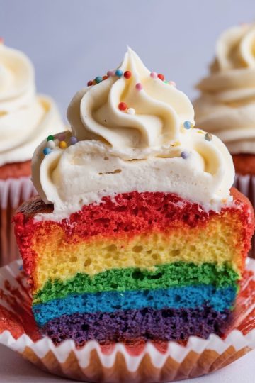 A detailed shot of a rainbow cupcake cut open, highlighting its vividly colored layers and topped with a swirl of vanilla frosting sprinkled with multi-colored confetti. The rainbow layers and smooth frosting swirl make this a visually appealing dessert, ideal for festive events or bright baking inspiration.