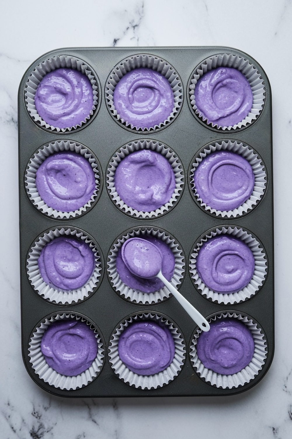  A muffin tin lined with paper cups, each filled with vibrant purple cupcake batter. A small spoon rests in one cup, showcasing the thick, creamy texture of the batter. This setup is ideal for colorful baking projects.
