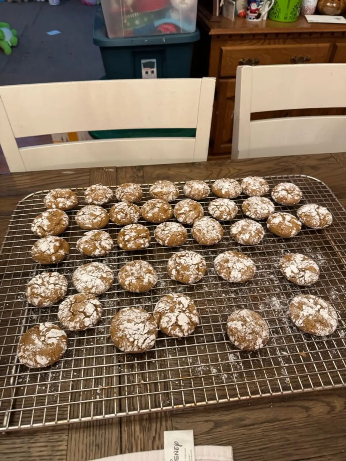 Freshly baked gingerbread crinkle cookies cooling on a wire rack, each cookie topped with a generous dusting of powdered sugar. Warm brown tones with cracked tops, arranged in neat rows on a wooden table.

