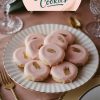 A single image displaying rose gold-glazed sandwich cookies elegantly arranged on a scalloped white plate with gold trim. The cookies feature a glossy pink glaze with delicate golden accents. The plate is styled on a blush pink tablecloth with crystal glassware and gold-toned cutlery, creating a sophisticated presentation.