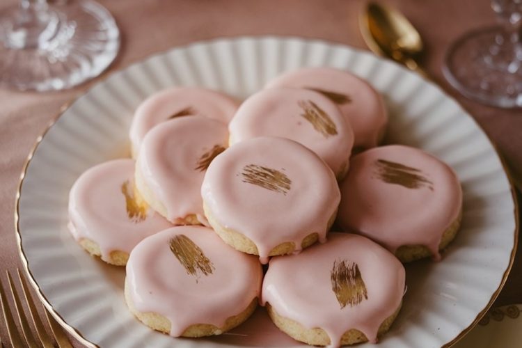 A single image displaying rose gold-glazed sandwich cookies elegantly arranged on a scalloped white plate with gold trim. The cookies feature a glossy pink glaze with delicate golden accents. The plate is styled on a blush pink tablecloth with crystal glassware and gold-toned cutlery, creating a sophisticated presentation.