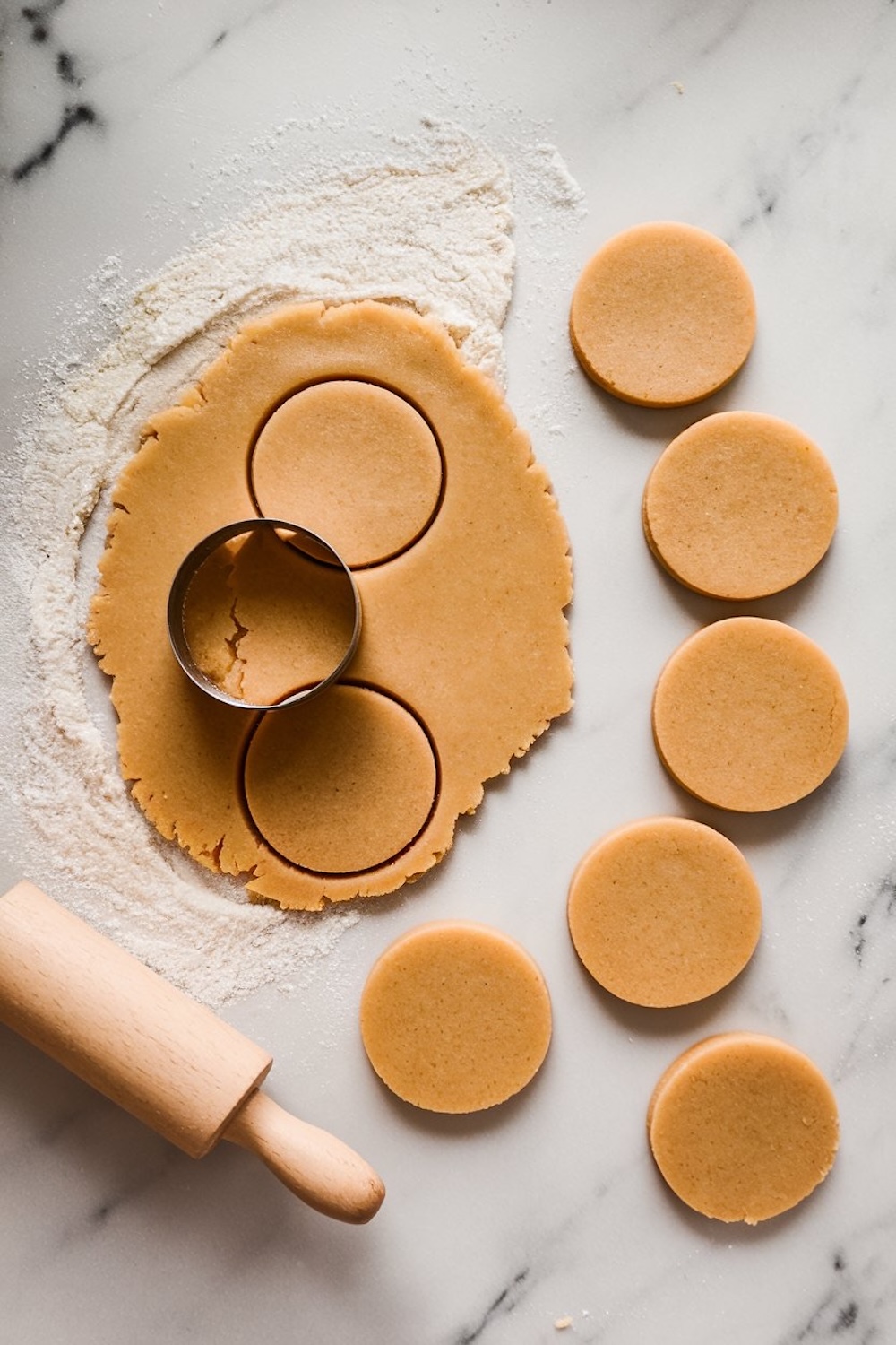 A single image showing raw cookie dough rolled out on a floured marble surface. Circular shapes are cut out with a round cutter, with a wooden rolling pin placed to the side. Unbaked cookie rounds are lined up next to the dough.