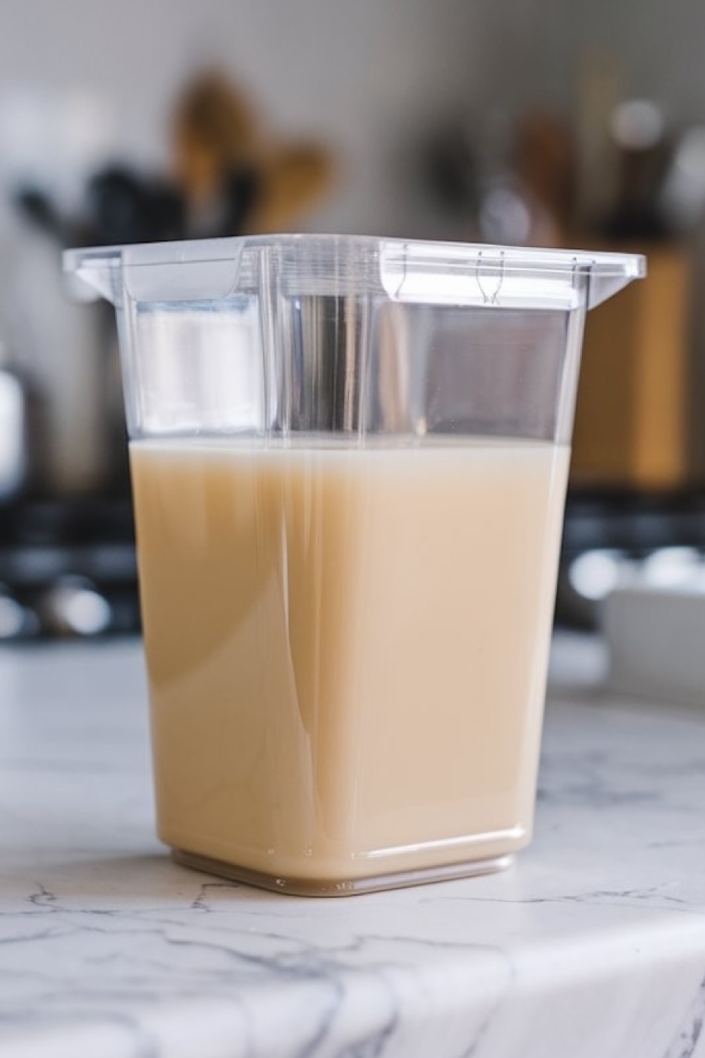 A clear, square-shaped container filled with creamy, beige-colored liquid a vanilla sorbet base, sits on a marble countertop in a home kitchen and about to get frozen
