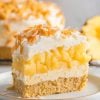 A single slice of pineapple coconut dump cake served on a white plate. Layers of creamy topping, golden pineapple, and a crumbly graham cracker crust are visible. Topped with a generous sprinkle of toasted coconut flakes, with a blurred whole cake and pineapple slice in the background.