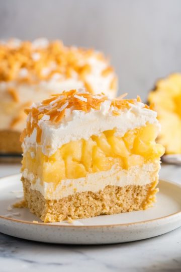 A single slice of pineapple coconut dump cake served on a white plate. Layers of creamy topping, golden pineapple, and a crumbly graham cracker crust are visible. Topped with a generous sprinkle of toasted coconut flakes, with a blurred whole cake and pineapple slice in the background.