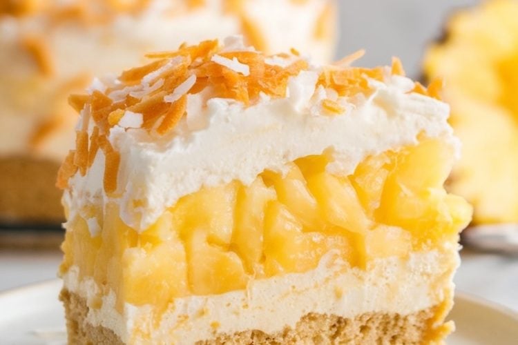 A single slice of pineapple coconut dump cake served on a white plate. Layers of creamy topping, golden pineapple, and a crumbly graham cracker crust are visible. Topped with a generous sprinkle of toasted coconut flakes, with a blurred whole cake and pineapple slice in the background.