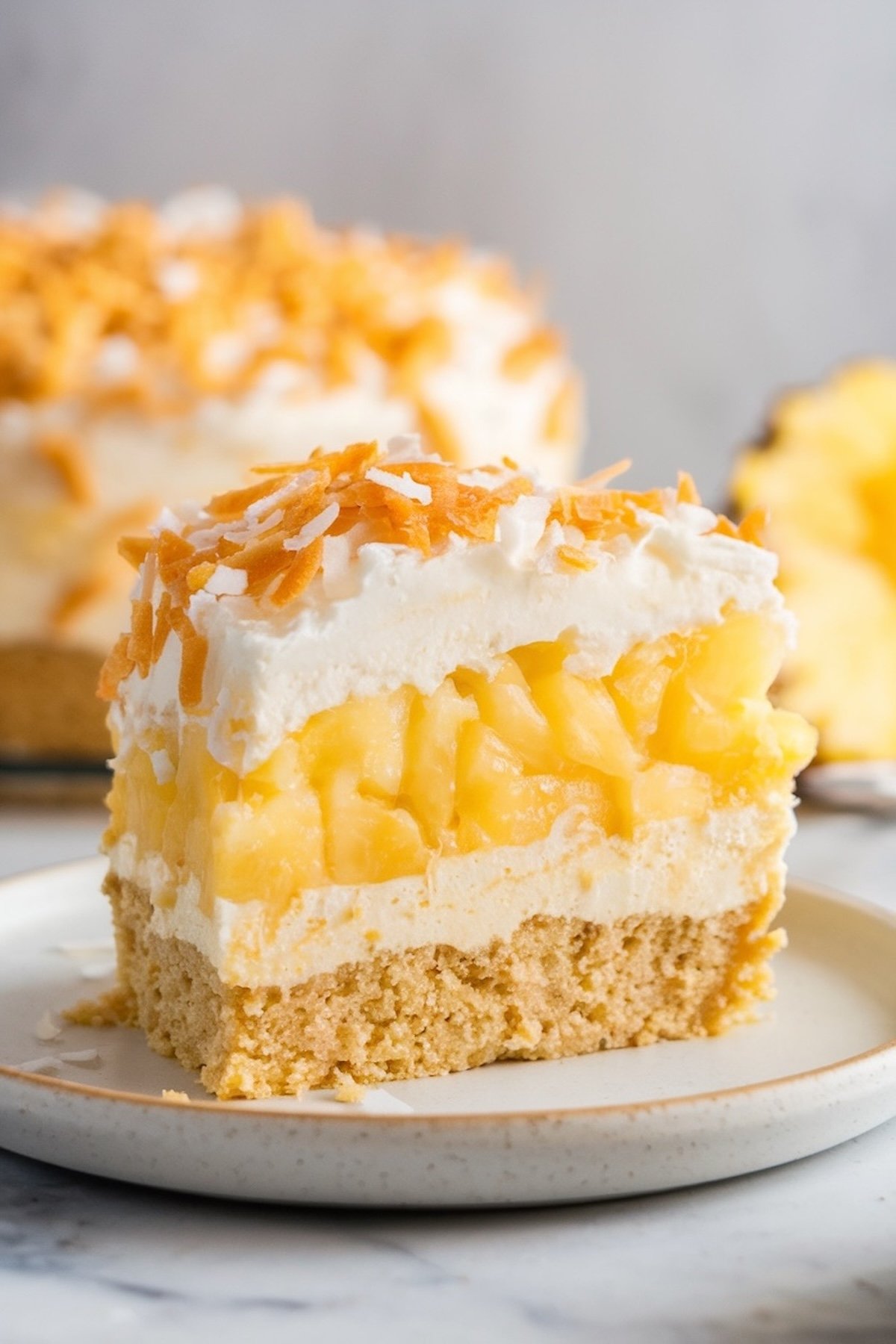 A single slice of pineapple coconut dump cake served on a white plate. Layers of creamy topping, golden pineapple, and a crumbly graham cracker crust are visible. Topped with a generous sprinkle of toasted coconut flakes, with a blurred whole cake and pineapple slice in the background.