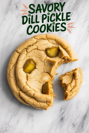 Overhead shot of multiple dill pickle cookies laid on a white marble surface, surrounded by flaky sea salt and a jar of pickles on a wooden board, highlighting the cookies’ golden texture and quirky garnish.