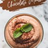 A single serving of avocado chocolate mousse dusted with cocoa powder and garnished with fresh mint leaves. The dessert sits in a glass, with text overlay reading “Avocado Chocolate Mousse” in bold, brushstroke-style font.