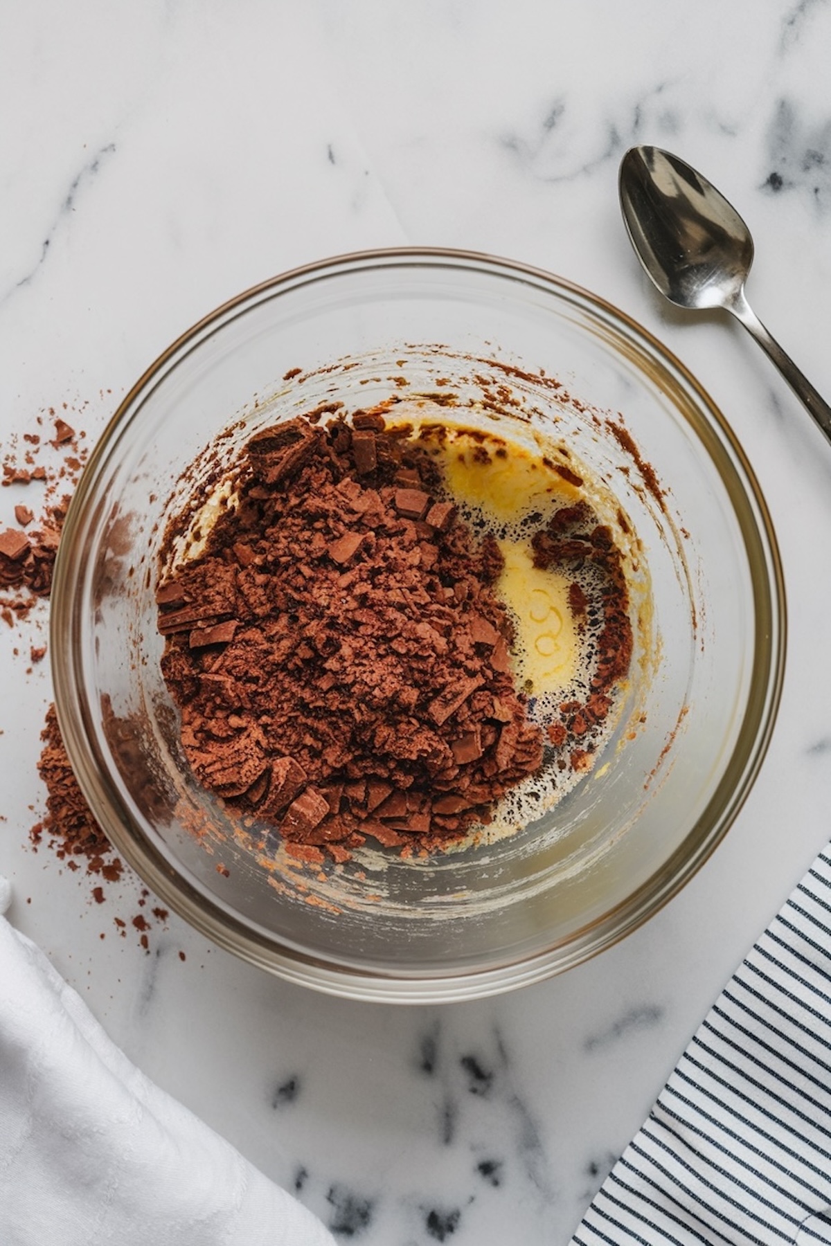 A mixing bowl with melted butter and chopped chocolate, beginning to combine into a chocolate mixture. A metal spoon rests beside the bowl on a striped kitchen towel.