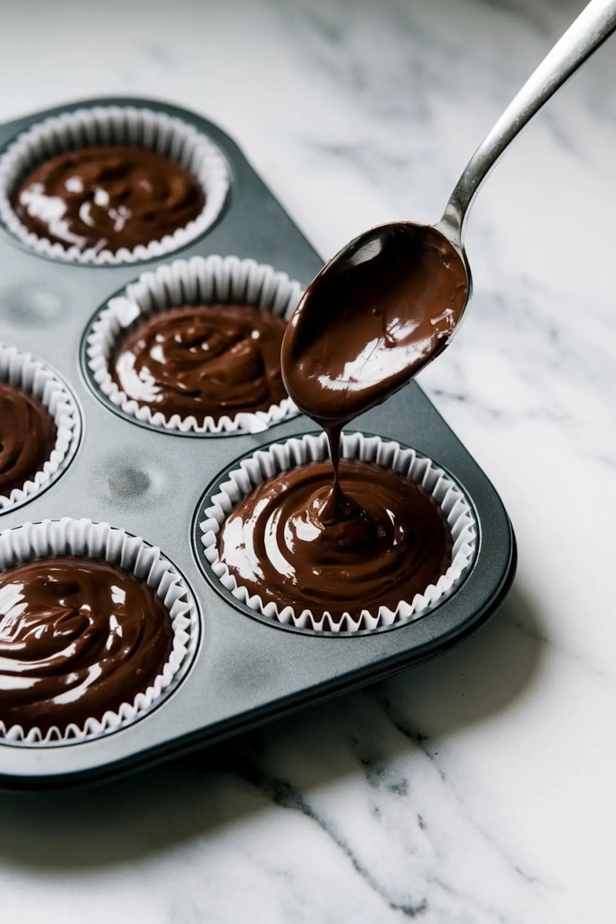 A close-up shot of chocolate cupcake batter being poured into white cupcake liners inside a muffin tin. The rich, glossy texture of the batter is highlighted against a marble countertop background.