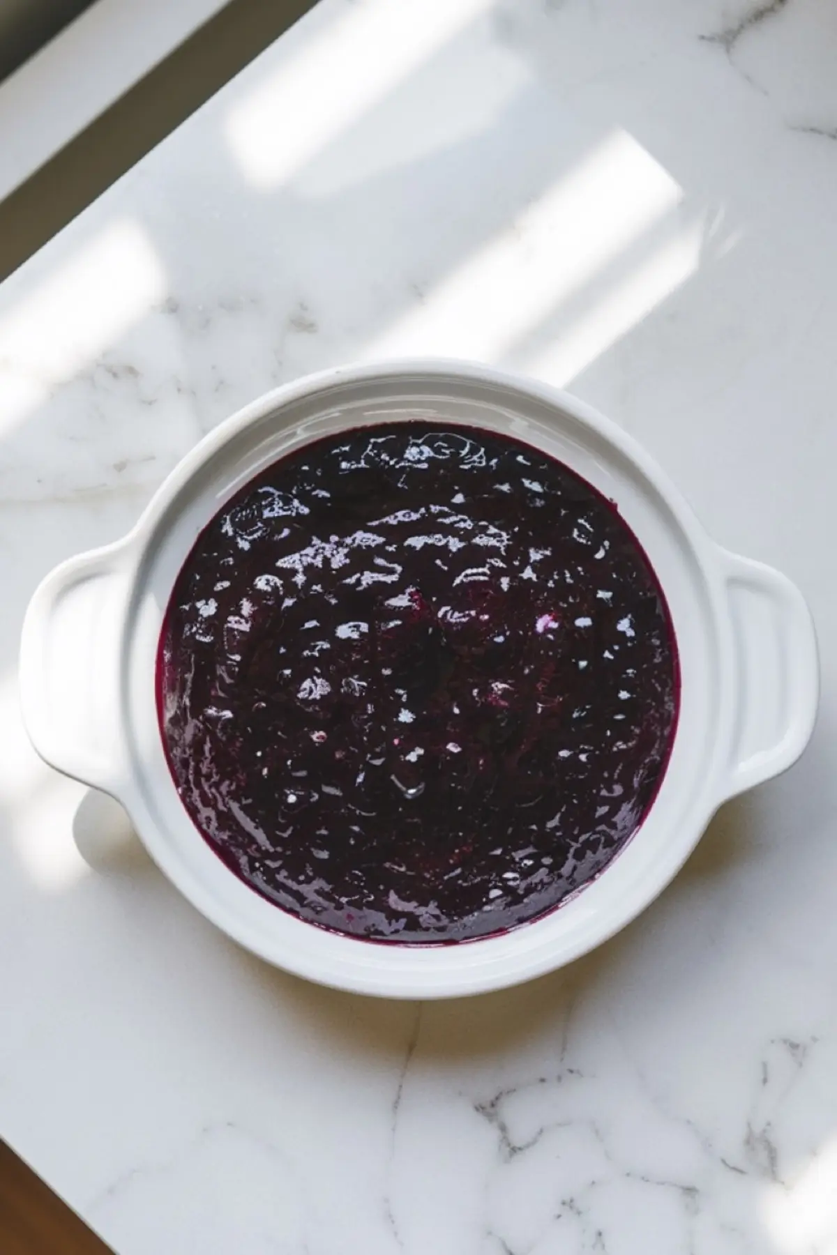Overhead shot of homemade blueberry filling in a white ceramic baking dish. The deep purple blueberry mixture has a glossy texture and sits on a marble countertop with soft natural lighting.
