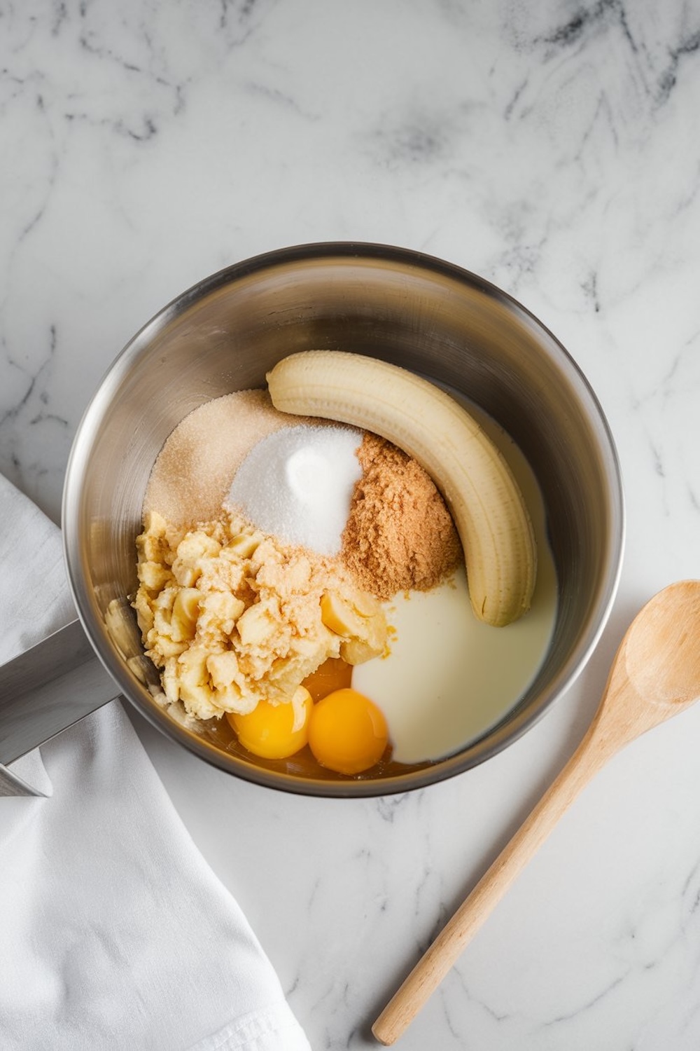 Mixing bowl with mashed bananas, whole bananas, eggs, sugar, brown sugar, and milk, ready to be combined for the blueberry banana bread recipe. A wooden spoon is placed beside the bowl.