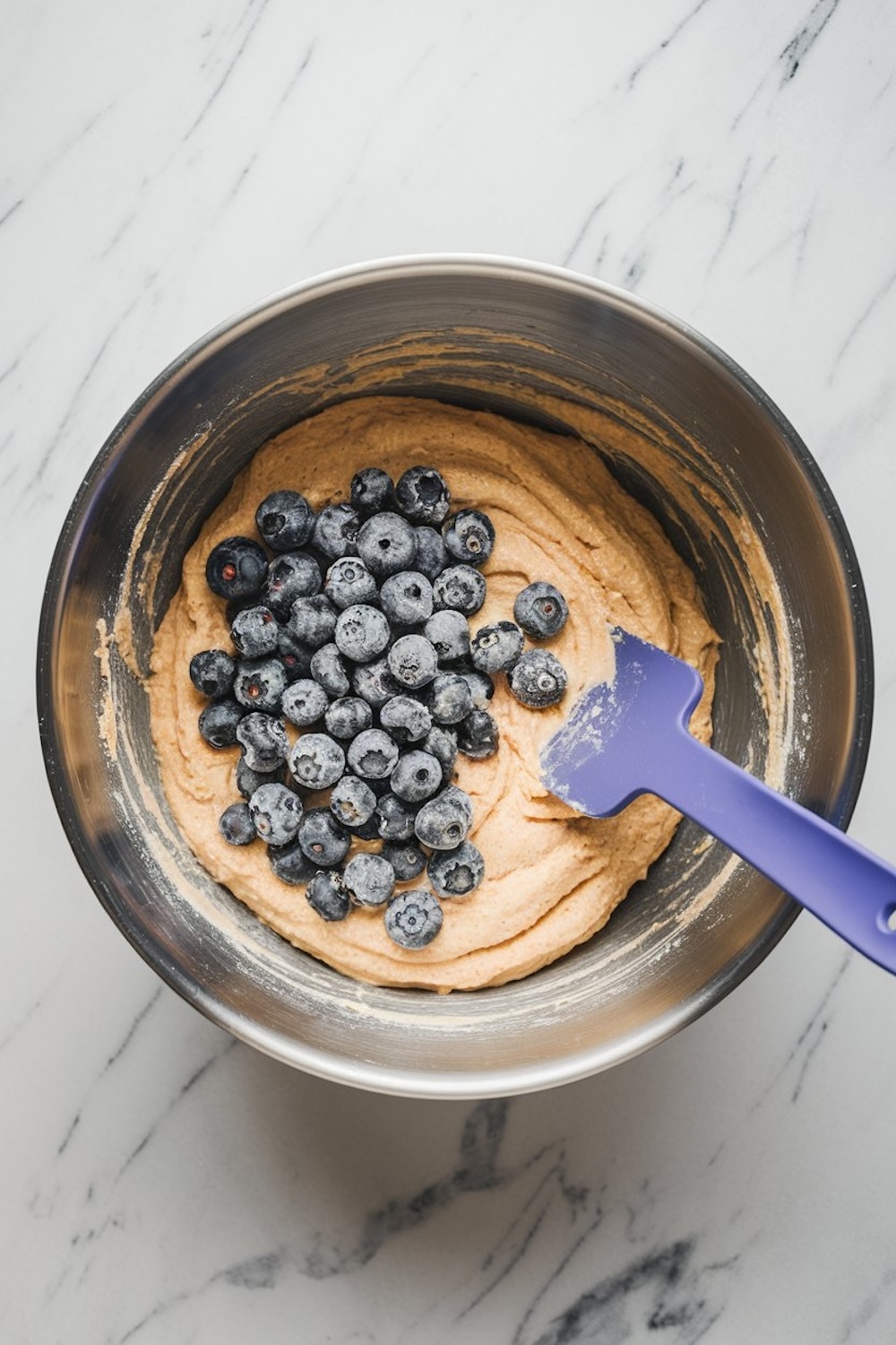 Mixing bowl containing blueberry banana bread batter, topped with fresh blueberries, and a blue spatula partially folding them into the mixture.