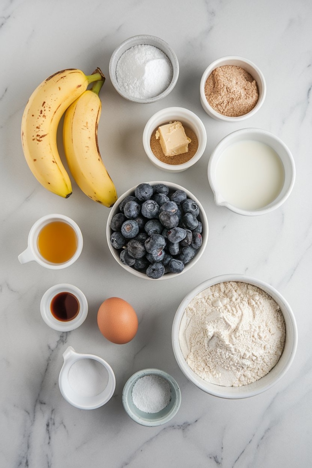 Flat-lay arrangement of ingredients for blueberry banana bread on a marble countertop. Includes bananas, fresh blueberries, flour, sugar, brown sugar, butter, egg, milk, vanilla extract, and baking soda, all neatly placed in bowls and cups.