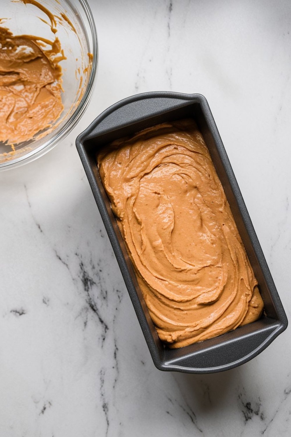 A loaf pan filled with smooth banana bread batter, placed next to a mixing bowl with remnants of batter, on a marble surface.