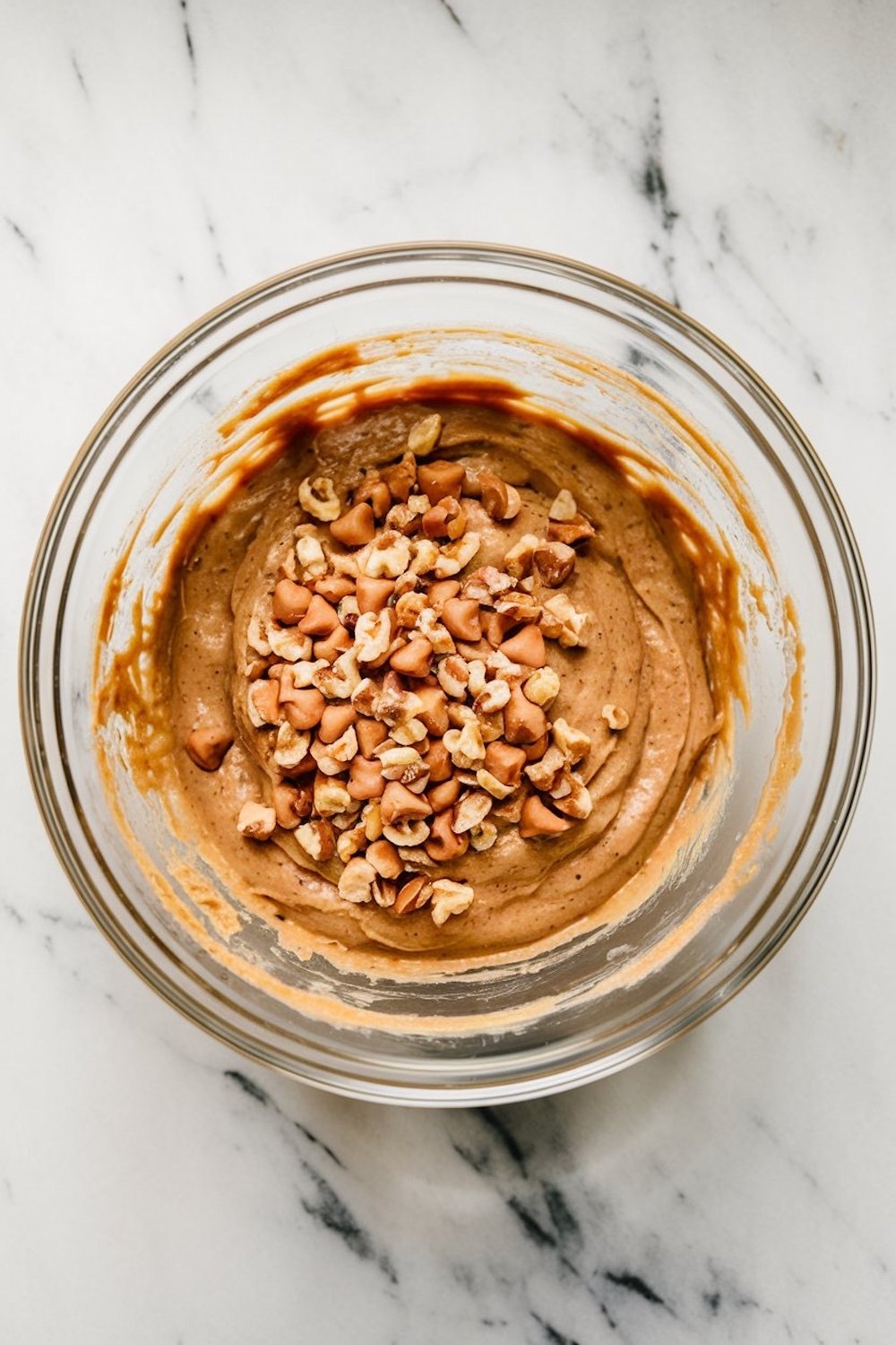 A mixing bowl with banana bread batter topped with chopped walnuts and butterscotch chips, ready for final mixing.