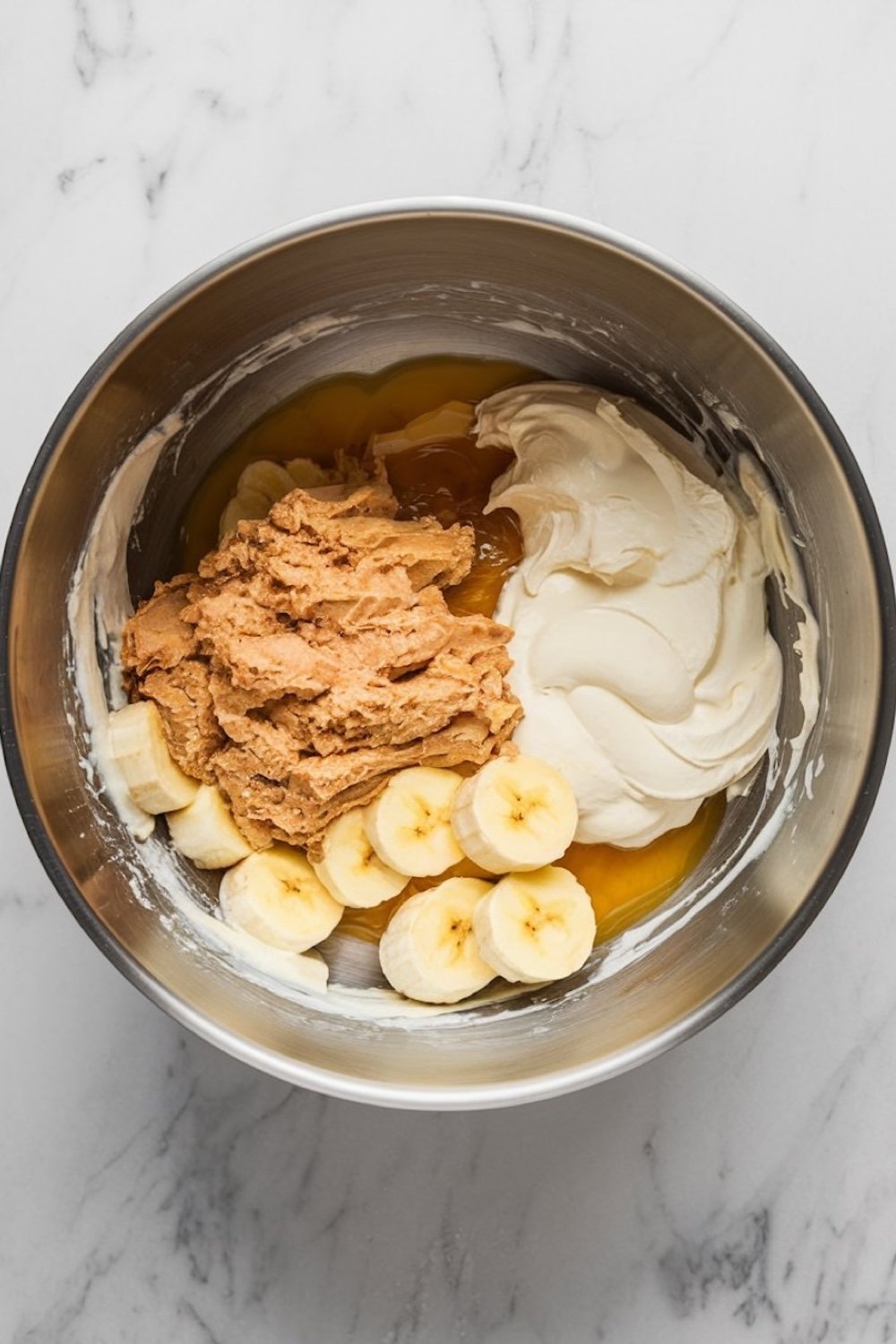 Ingredients for caramel pecan banana bread in a mixing bowl, including sliced bananas, peanut butter, yogurt, and caramel sauce, layered and ready to be blended. The metal bowl rests on a white marble surface.