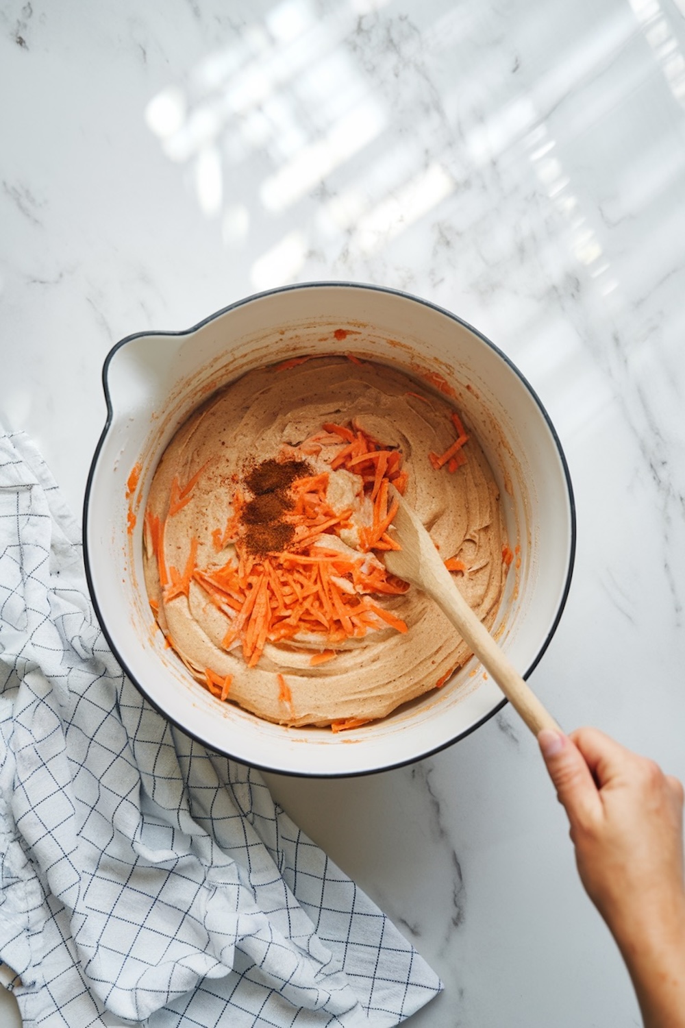 A mixing bowl with carrot cake batter. The batter is light brown with visible shredded carrots and a sprinkling of spices on top, being stirred with a wooden spoon.