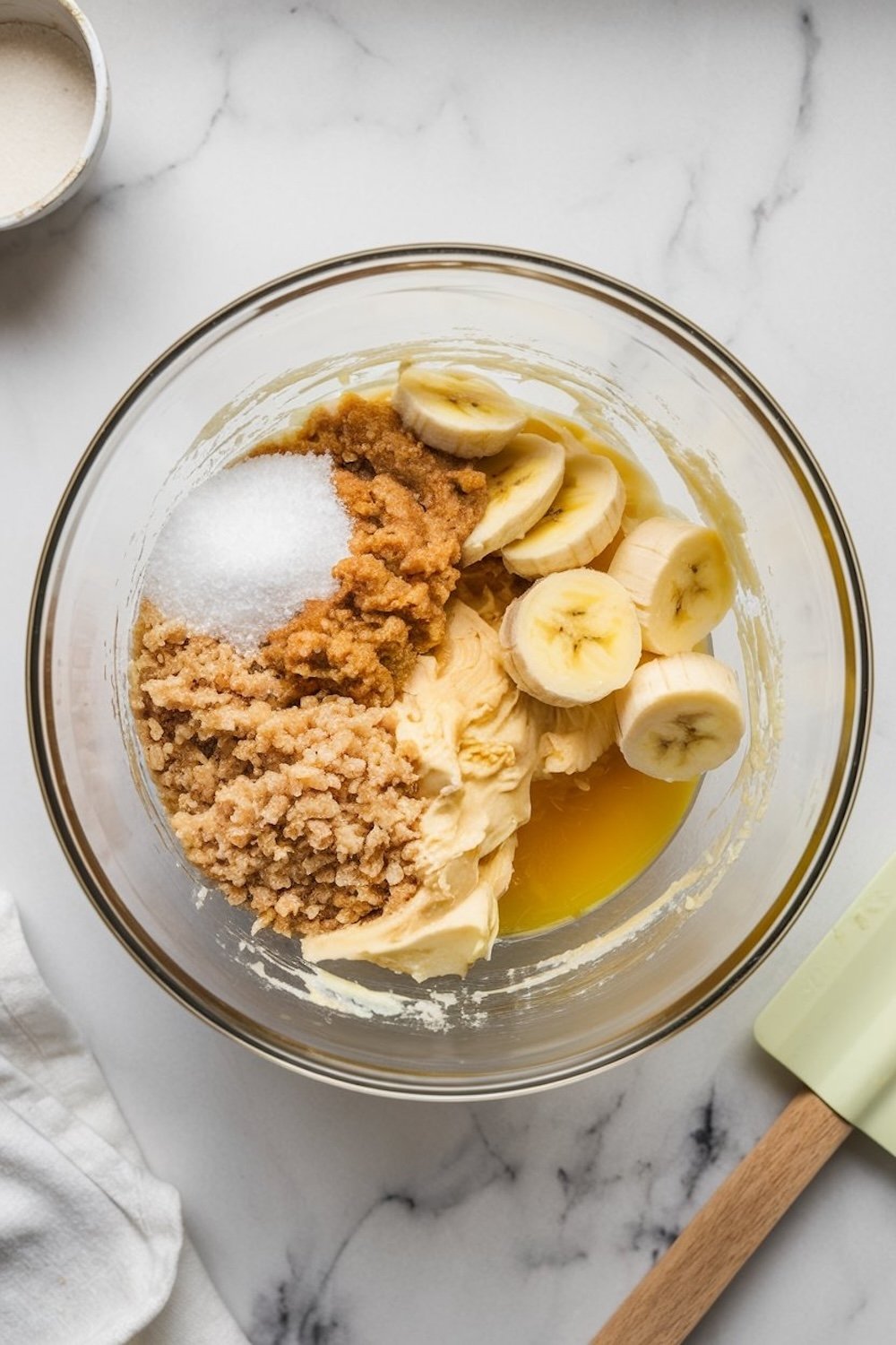Glass mixing bowl containing sliced bananas, brown sugar, granulated sugar, melted butter, and cream cheese on a marble surface. Ingredients are partially blended, ready for mixing. A spatula and additional bowls are visible nearby.