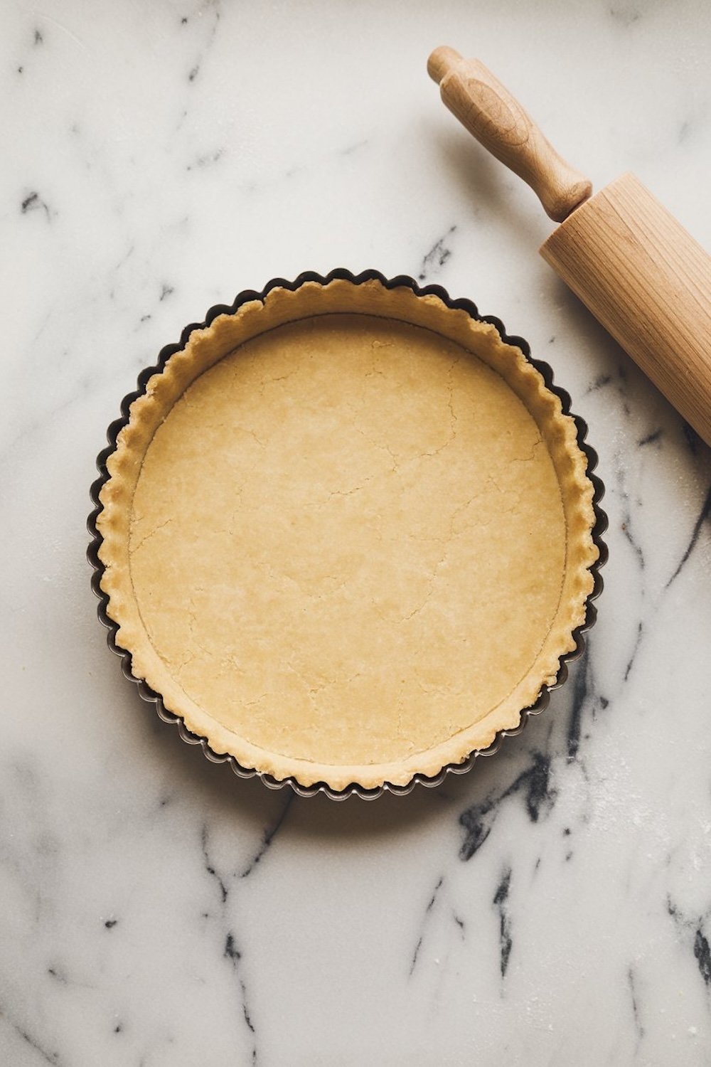 A pre-baked tart shell in a fluted tart pan on a marble surface with a wooden rolling pin placed beside it.